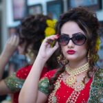 Beautiful South Asian bride in traditional red attire, adorned with jewelry and sunglasses, posing indoors.